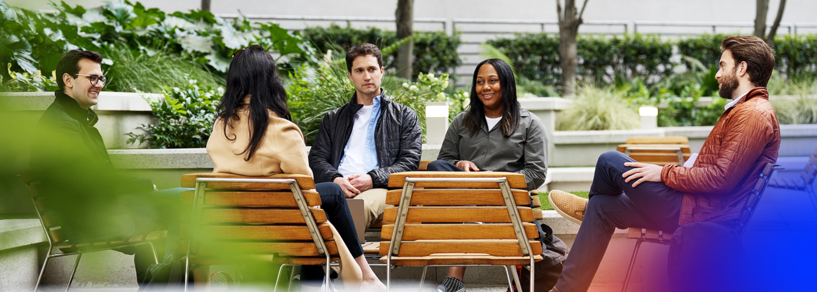 A group of five diverse young professionals sits in a semi-circle on wooden outdoor chairs in a lush, landscaped courtyard. They appear to be engaged in a casual meeting or collaborative discussion. The setting features modern concrete planters filled with greenery and soft outdoor lighting, creating a bright and relaxed professional atmosphere.