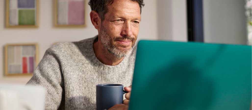 Image of a man inside an office space holding a cup of coffee while looking at a laptop.