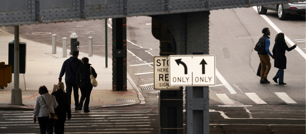 Image of a city street that overlooks under a bridge of groups of people walking across crosswalks.