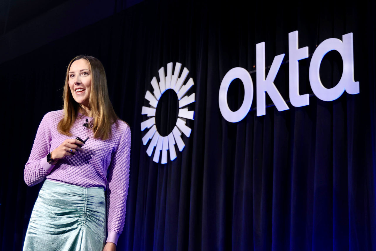 Charlotte Wylie speaking on a stage in front of a large white Okta logo and dark curtains.