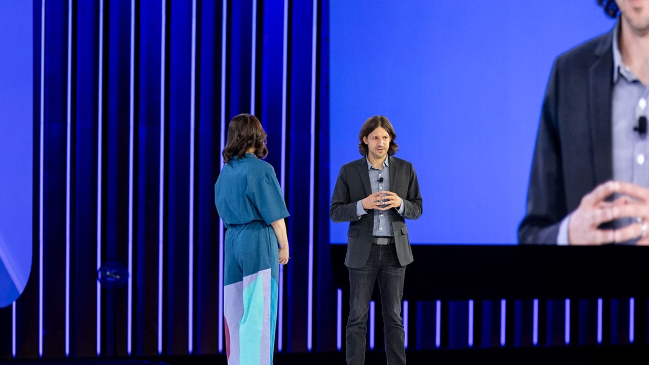 A man and a woman are on stage at the Oktane conference, standing in front of a large blue screen with vertical light strips.