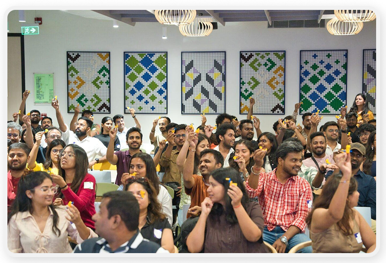 A large group of people sit closely together in a modern indoor venue, many of them raising their hands or holding up lego objects. 