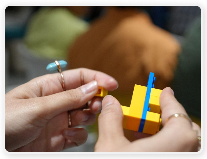 A close-up view shows a person assembling small yellow and blue plastic building blocks. 